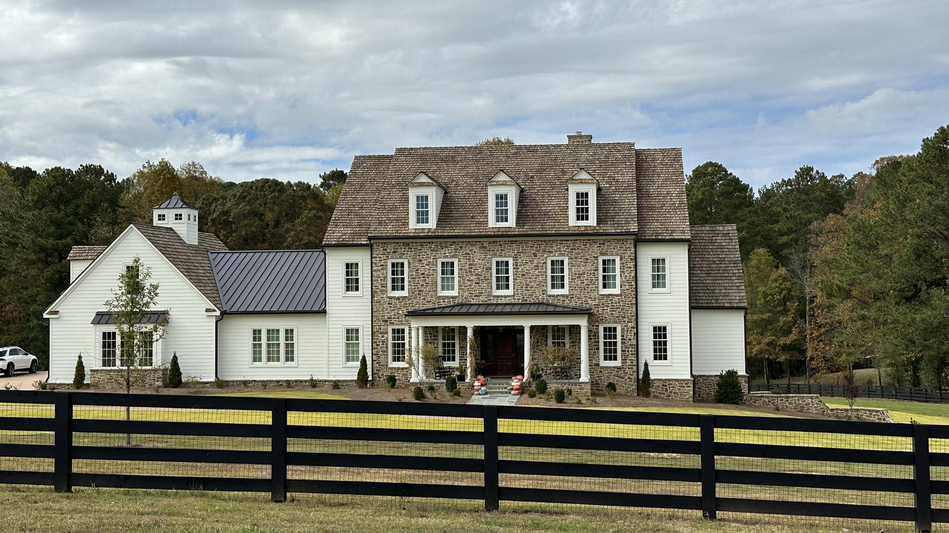 Stone and lap siding country home with cedar shake roof behind a black board fence